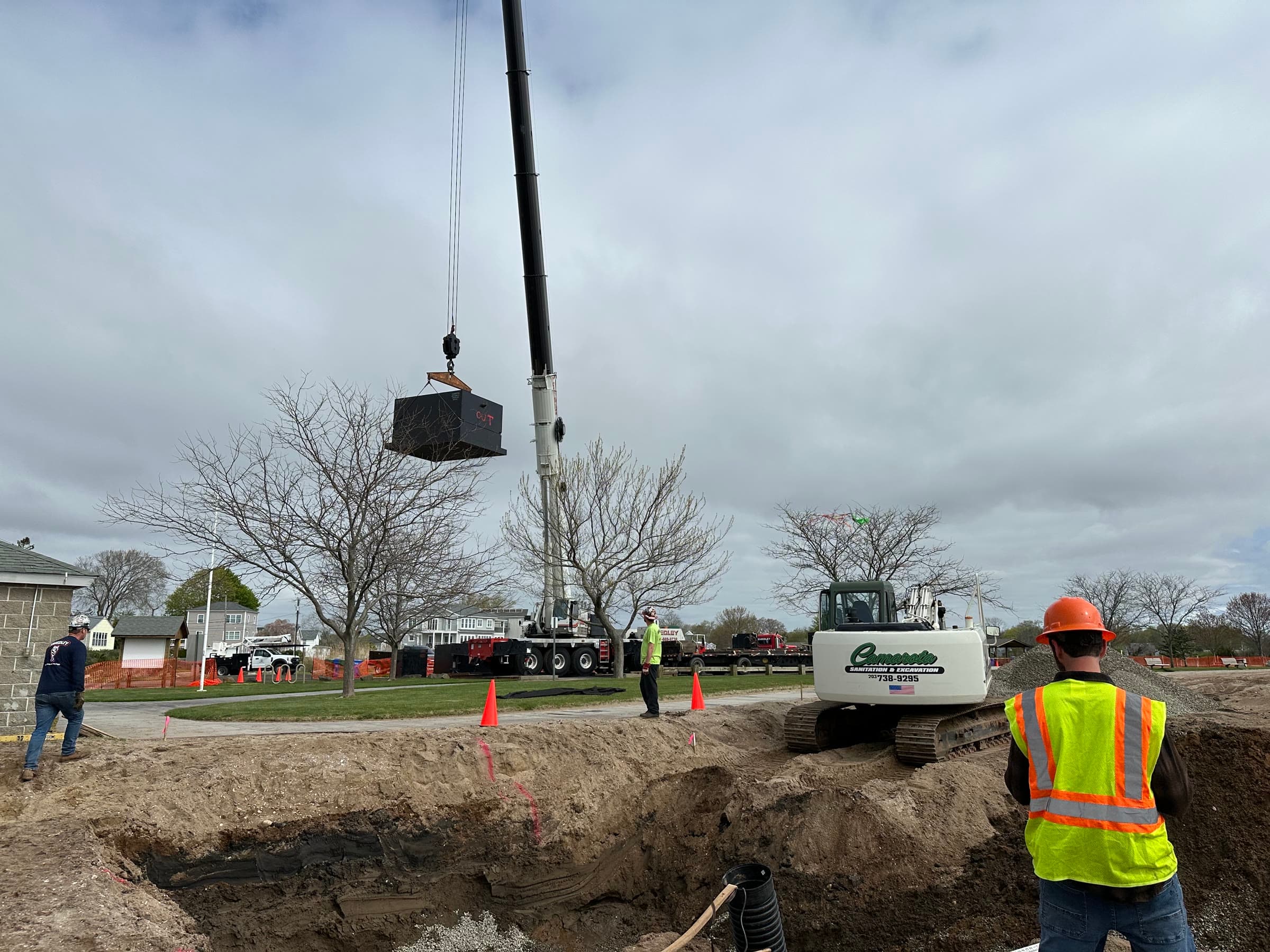 Concrete septic tank on a crane above the excavation, with a Camarota excavator and crew on site.